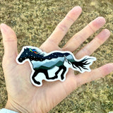 Hand holding a sticker of a horse with a starry sky design against a grassy background.  Horse-shaped sticker with a scenic design held in front of a natural landscape.  Waterproof vinyl sticker, Horse sticker, Vinyl decal, Laptop sticker, Water bottle sticker, Horse silhouette art, Die-cut sticker, Celestial sticker, Wildlife sticker, horse silhouette sticker, mountain sticker, feather sticker, watercolor feather sticker